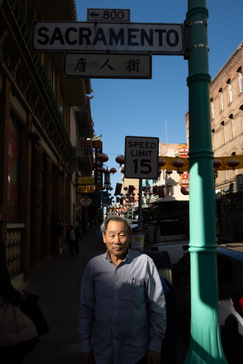 An Asian American man stands on a city corner under street signs, one of which reads "Sacramento."