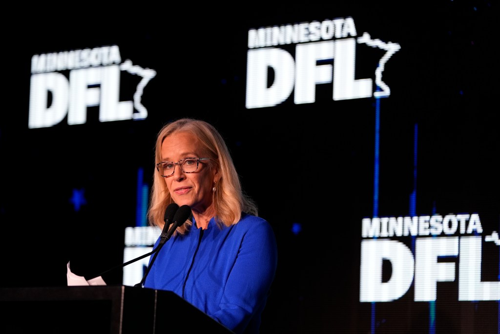A blonde woman in a bright blue suit speaks at a podium with the Minnesota DFL logo in the background.