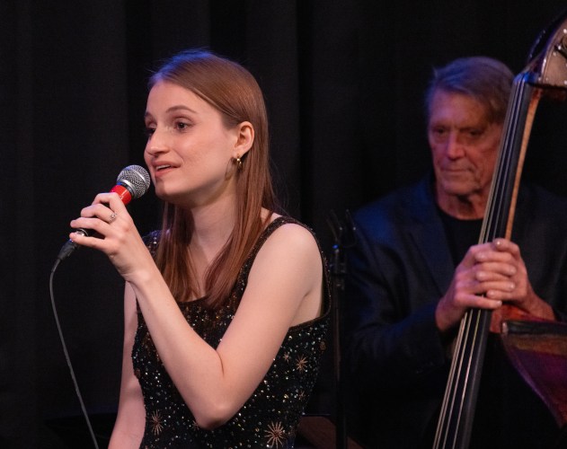A woman with long hair sings into a mic in front of a man playing upright bass.