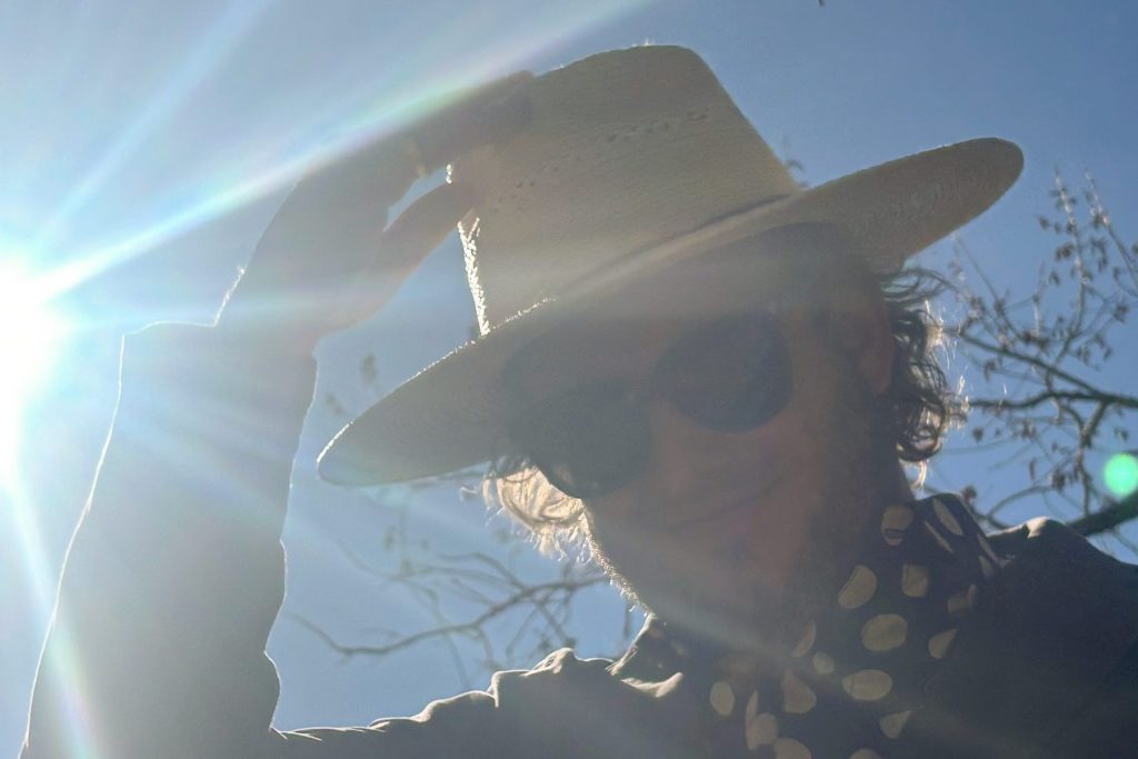 Singer Justin Vernon tipping a hat, sunshine behind him