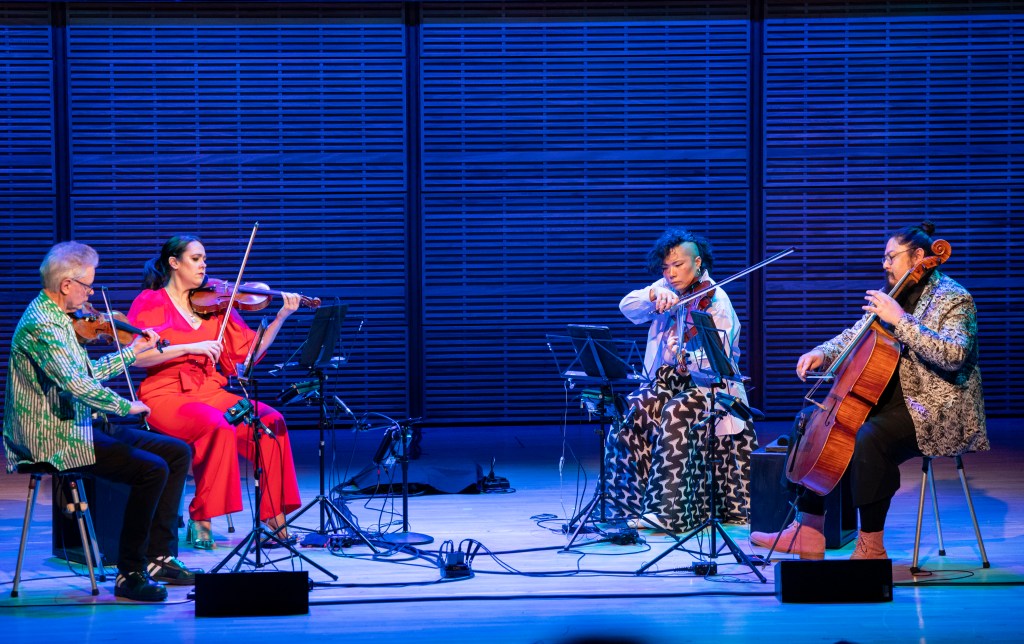 Four people playing stringed instruments, the Kronos Quartet, sit on a stage in a half circle.