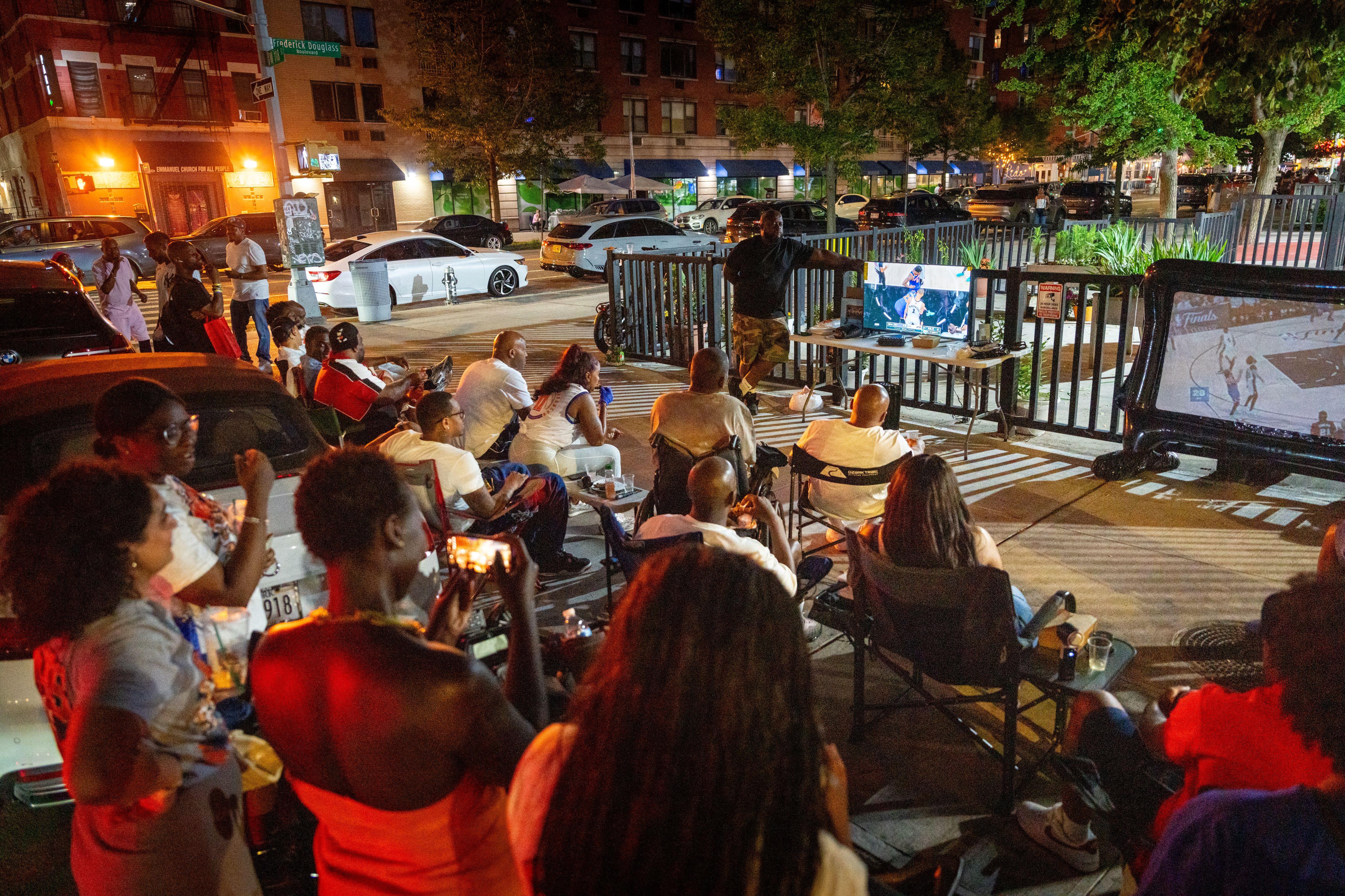 People gather on a sidewalk and beside cars to watch a Knicks game on three screens.