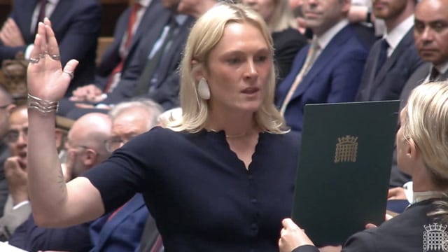 Lara Bird with her right hand in the air in Parliament and her three middle fingers appear to be crossed