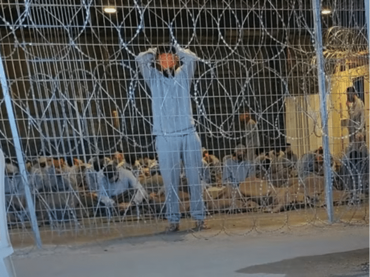 A leaked photograph of the Sde Teiman detention facility shows a blindfolded Palestinian man with his arms above his head behind a metal wire fence in 2024. Knesset politicians defeated a bill to ban visits from the Red Cross.