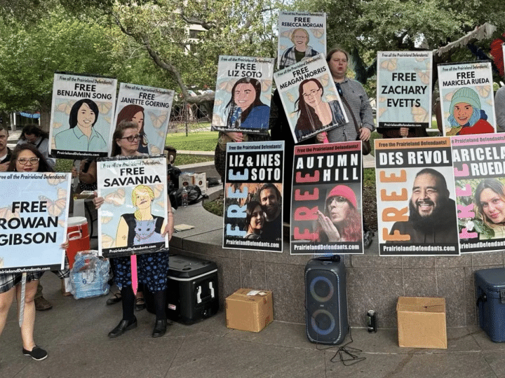 Posters with 'Free XYZ' line a wall and one woman holds one saying 'Free Rowan Gibson'. They are part of the protestors jailed for demonstrating outside an ICE centre