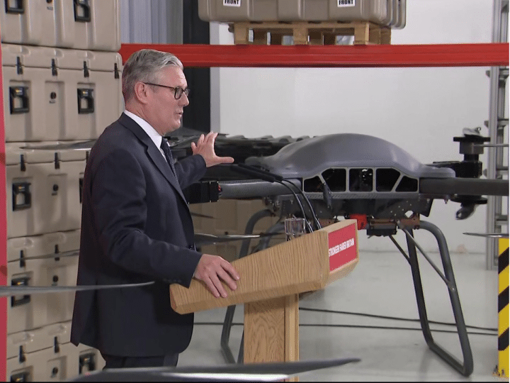 Keir Starmer touching the top of a drone inside a company factory, speaking to a crowd at a wooden podium. We cannot see the crowd.