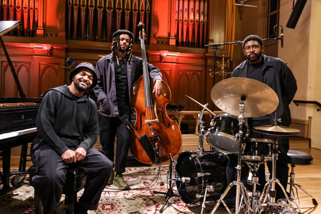 Three men stand by an upright bass and drumset.