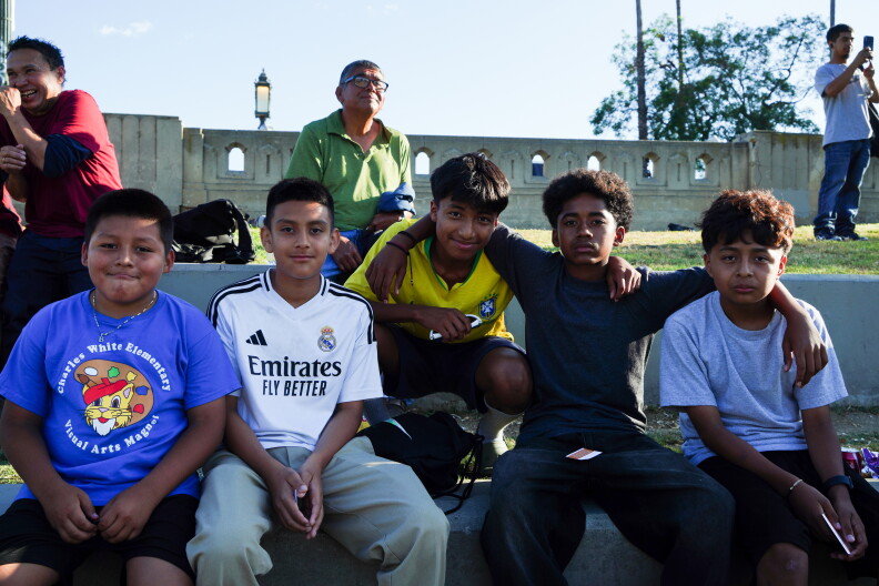 Five boys sit on a ledge at a park. One of them wears a blue shirt that reads "Charles White Elementary Visual Arts Magnet." Two of the other boys wear soccer jerseys. 