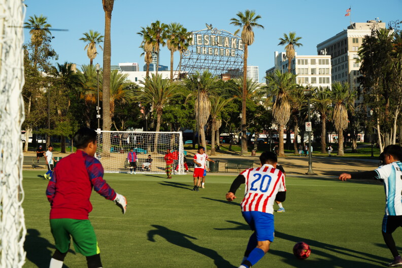 About a dozen men in different-colored uniforms play soccer at a palm tree-lined park. A sign that reads "Westlake Theatre" can be seen against a bright blue sky, surrounded by multi-storied buildings. 