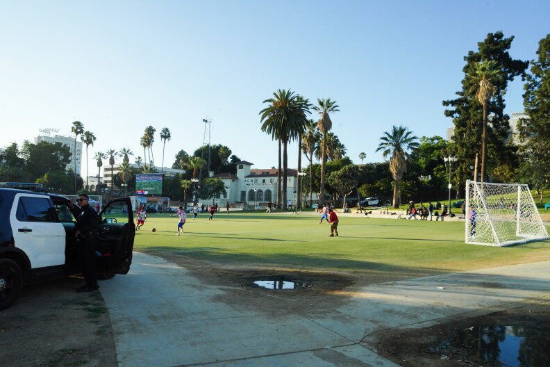 Young men play soccer in a park while a police officer watches on from outside a patrol car.