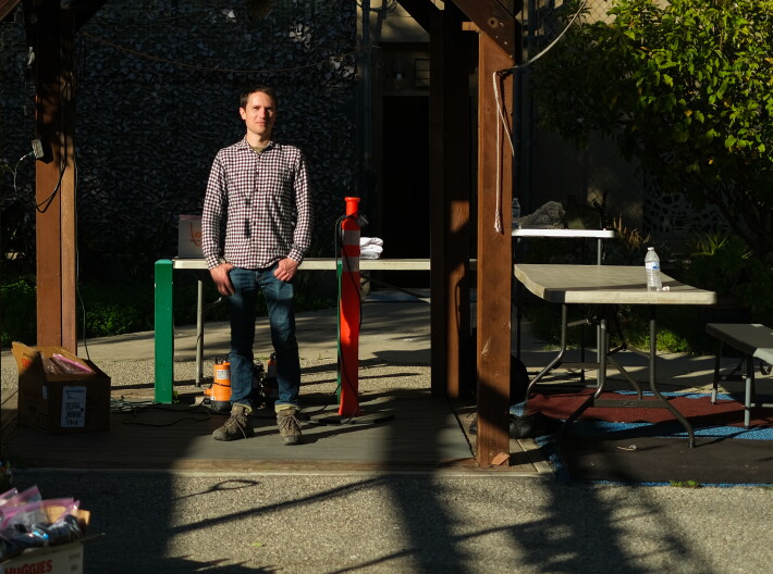 A man in a checkered shirt and jeans stands in front of two tables.