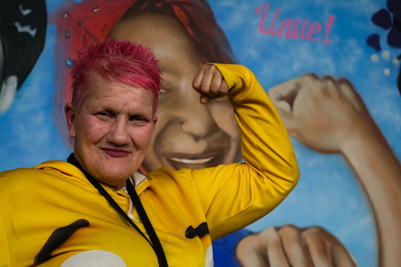 A woman with short, pink hair stands with her left arm flexed in front of a mural.