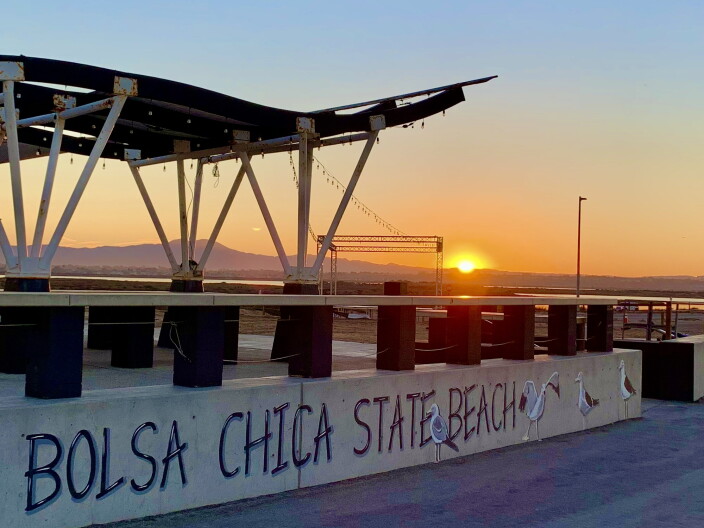 The sun rises in the distance while in the foreground, there's a concrete wall that says Bolsa Chica State Beach with paintings of gulls.