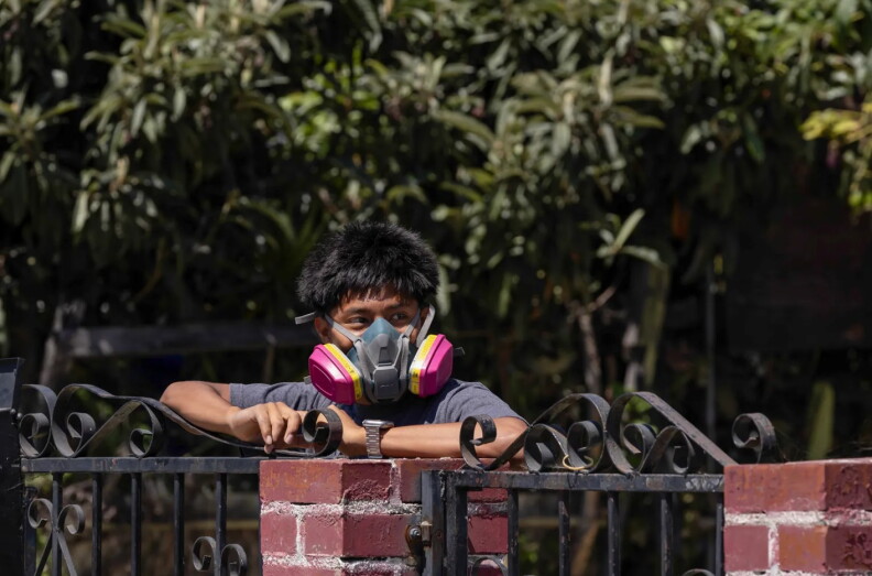 a child wears a gas mask and leans up against a fence with some bushes and trees in the background