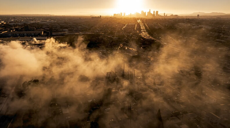 smoke spreads across a charred landscape as the sun sets behind a distant cityscape.