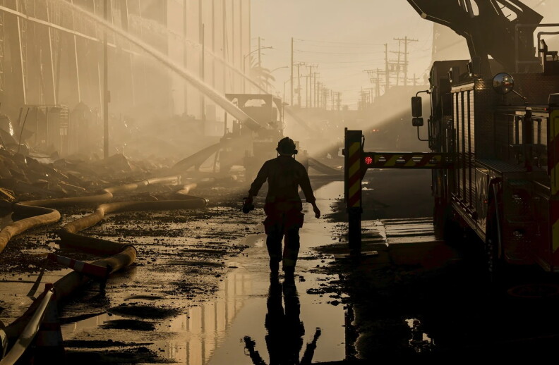 a man in a helmet walks past a lot of debris and fire equipment as hoses spray water