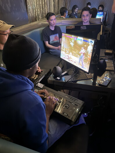 Person plays video game on monitor with arcade stick, two onlookers in background.