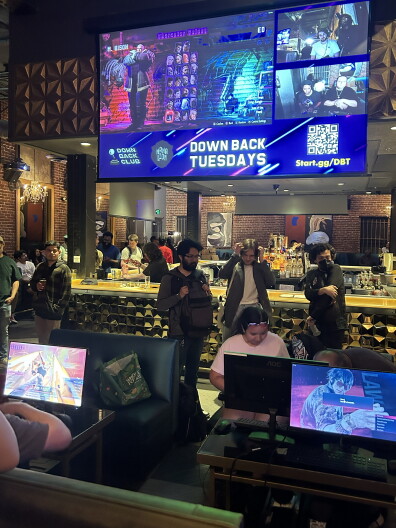 Gamers compete at a bar with a large screen showing 'Down Back Tuesdays' and a video game tournament display.