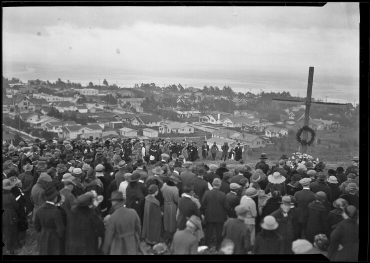 A wide archival view of a large crowd of people standing on a hill overlooking a neighorhood full of homes. A cross with a wreath can be seen toward the right in the middle of the crowd.