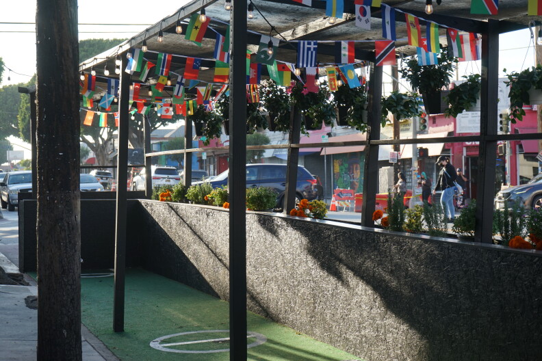 An empty patio painted in black sits empty. A string of international flaps are decorated above the roof of the structure with string lights. People in the distance are wearing face masks. 