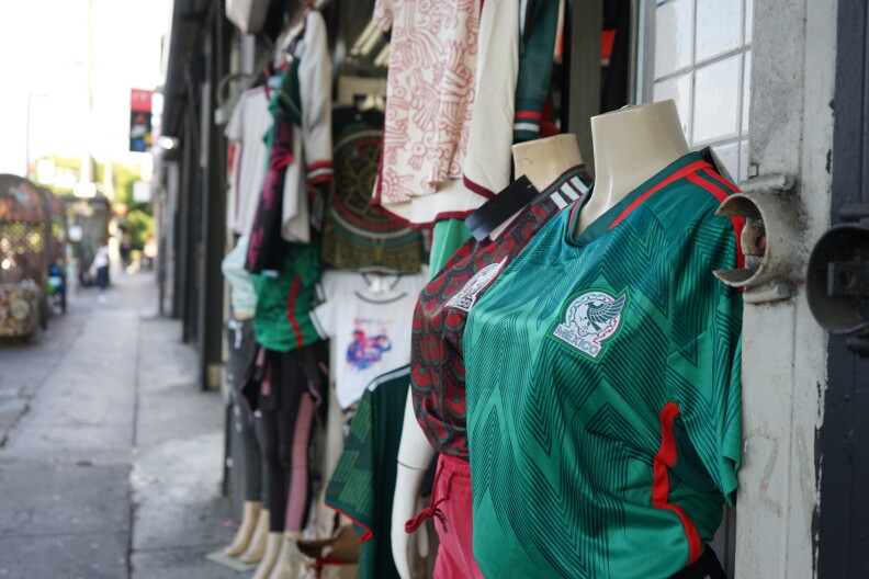 Mannequins wear a green and red jersey. A patch on the corner reads "Mexico. The mannequin is standing on the side of an empty sidewalk and surrounded by other shirts. 