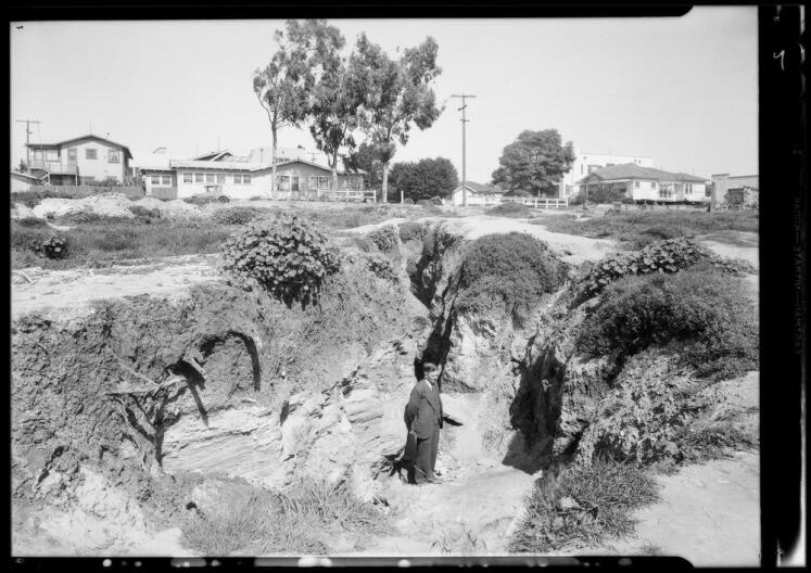A black and white archival of a man in a suit standing in a large ditch between two sections of land. The foundation he's standing on is much lower than the two sides and you can see the rock layers and roots sticking out of them. Homes are in the background.