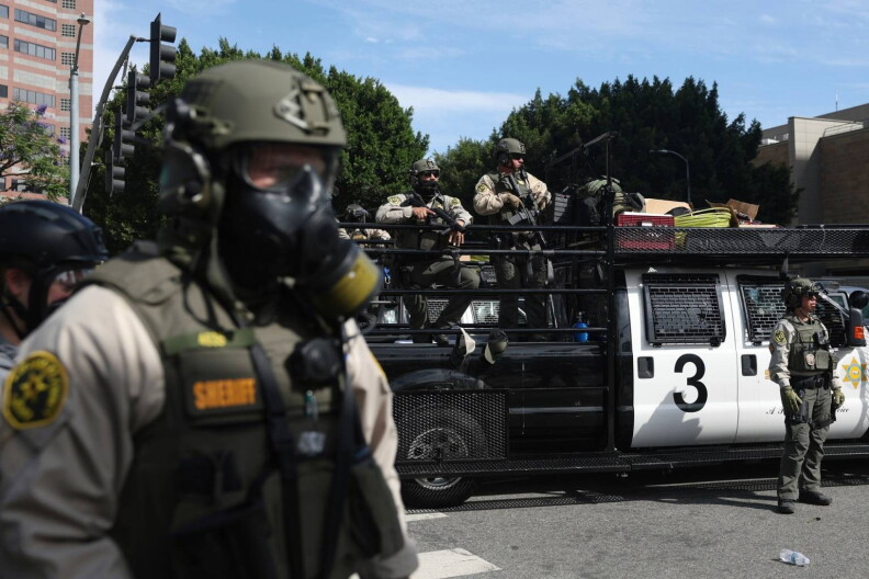 Armed officers wearing bullet proof vests, helmets and gas masks stand in front of and on top of a black and white truck with the number "3" on it.