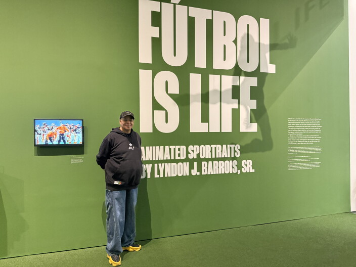 A man wearing a black hooded sweatshirt stands in front of a green wall with white text on it that reads: 'Futbol is Life'