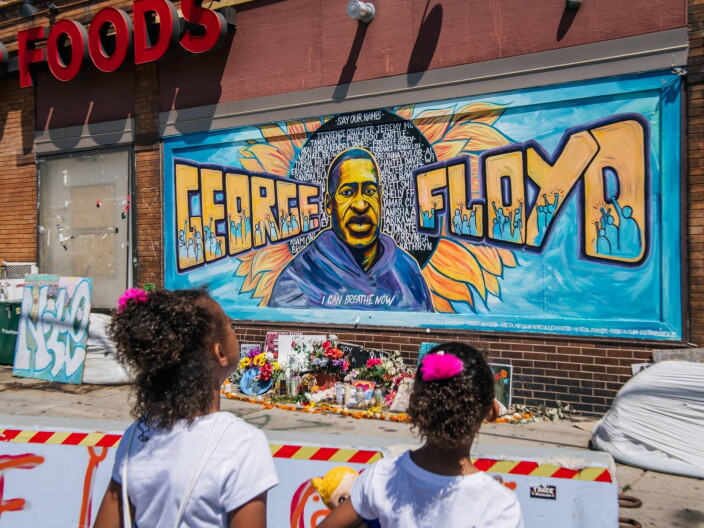 Two young girls with pink bows in their hair are pictured from behind stand in front of a mural. The mural features a man in a blue sweatshirt in the middle, with the name "George Floyd" painted in large orange letters.