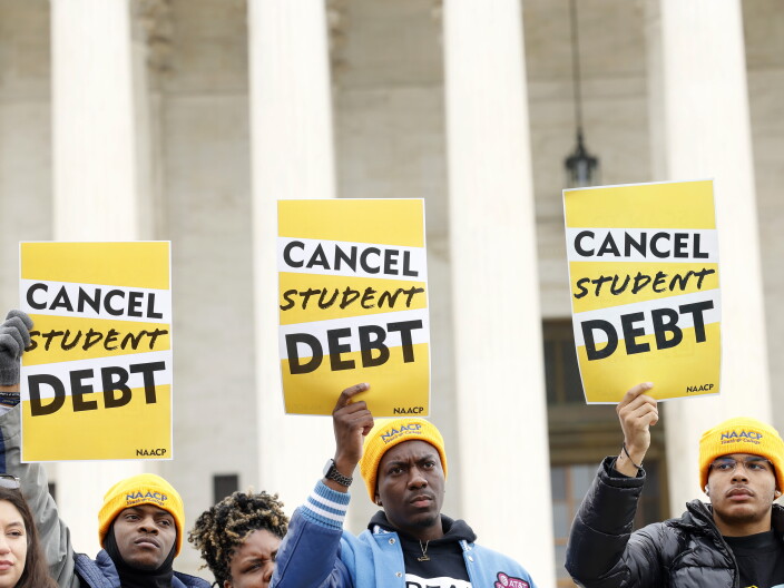 A group of young people stand in front of a building holding up signs that say "Cancel student debt."