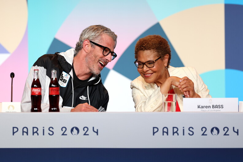 A man wearing glasses and a jacket that has a patch that reads "LA28". He leans in to speak to the woman on his left who is leaning in to hear him. They sit behind a desk that reads "Paris 2024."
