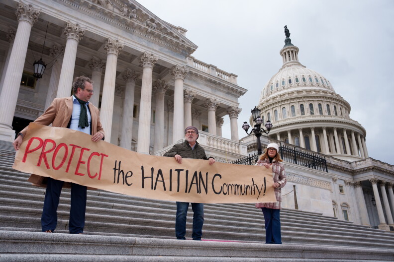 Three people stand on the steps of a stone building. They're each holding a sign that reads "Protect the Haitian Community!"