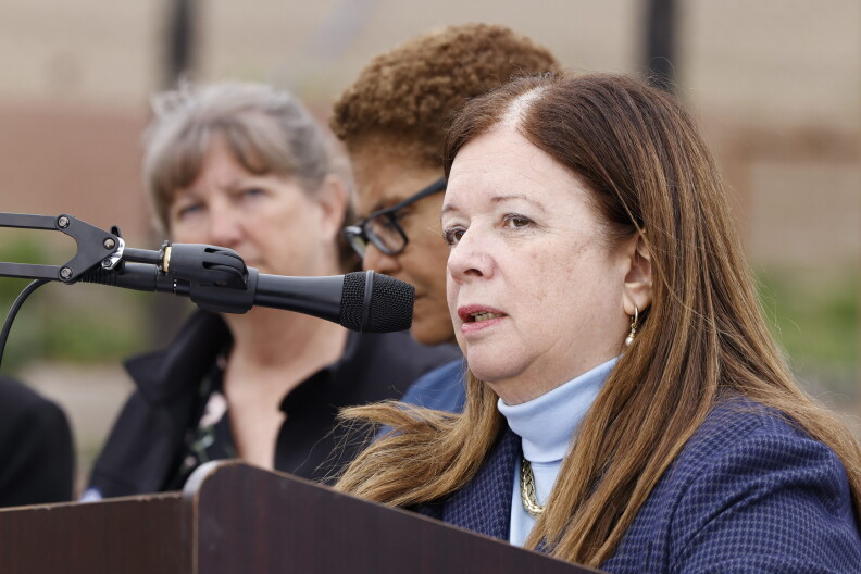 A woman with brown hair past her shoulders is speaking into a microphone affixed to a podium. She's wearing a light blue turtleneck under a navy blue checkered jacket and small earrings. Two other women can be seen standing behind her on the left.