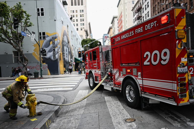 A red fire engine is parked in a street intersection. A firehouse attached to the engine is connected to a fire hydrant on the sidewalk. A firefighter stands beside the hydrant on the sidewalk.