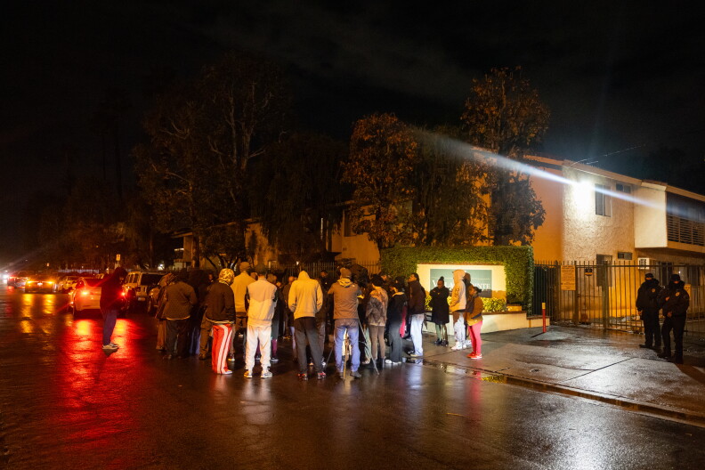 People gather in a tight circles outside a gated apartment complex. Men in uniforms with their faces partially covered are at the far right of the image.