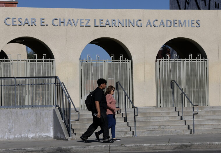 A young man with medium dark skin tone wearing all black, including a backpack, walks next to a woman with medium skin tone in a pink shirt. The letters on the building behind them read Cesar E. Chavez Learning Academies. 
