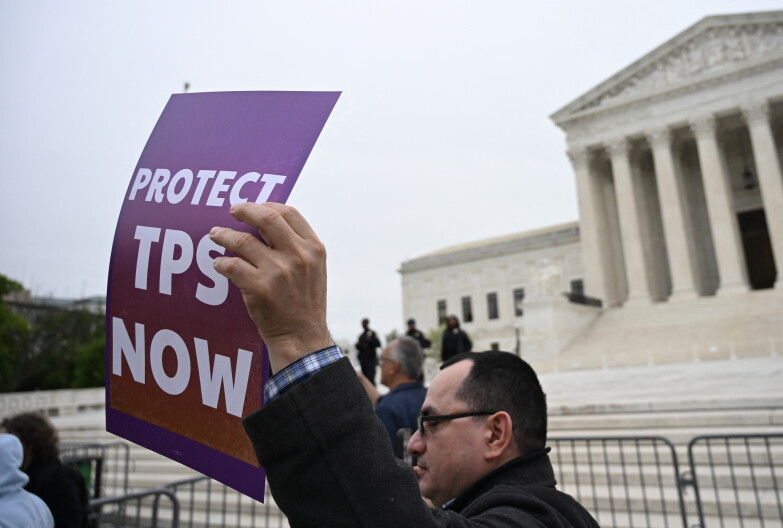 A man holds a sign that reads "Protect TPS Now". He stands outside a stone building.