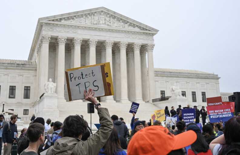 A person holds a sign that reads "Protect TPS" as they stand in a crowd. A large stone building is in the distance.