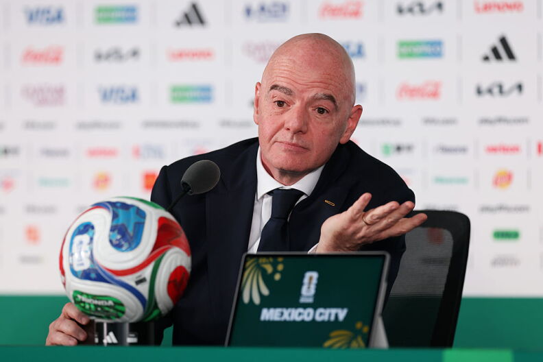 A man in a dark suit and tie sits at a desk with a podium mic and a blue, red, white and green soccer ball sitting in front of him. He's arching eyebrows and raising his hand up. 