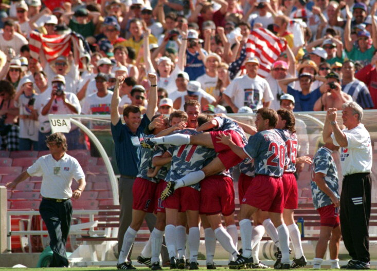 A group of male soccer players wearing red, white and blue celebrate on a green soccer pitch. A man in the middle is jumping into the group. 