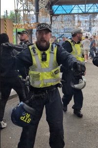 A police officer screaming at the camera. He is at the entrance to Brighton Train Station and he is brandishing his baton.