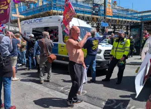 A bald man with his shirt off giving a thumbs up to the antifascist crowd. He is wearing black joggers and shoes. Behind him in the police cordon with police vans. All around him are other people.