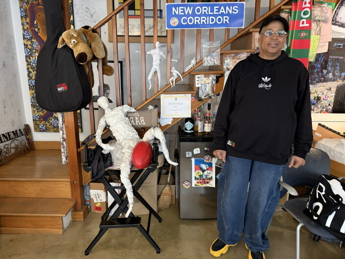 A black man wearing glasses standing in the middle of a room with a lot of sculptures and art.
