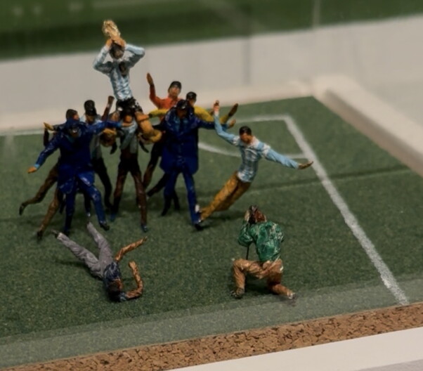A tiny scene of miniatures on a soccer field. A soccer player is hoisted above the heads of other players.