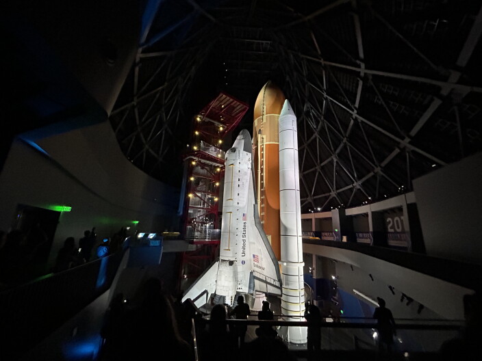 A space shuttle in its launch position stands in the middle of a museum exhibit. 