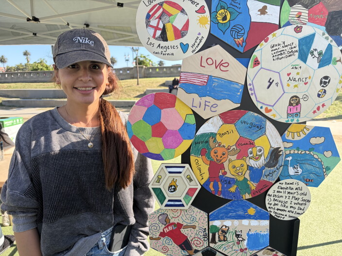 A woman with medium-light skin tone and long brown hair smiles gently, clad in a gray long-sleeved shirt and matching baseball cap. She stands next to a display of wooden circles and hexagons, decorated in bright colors by children. 