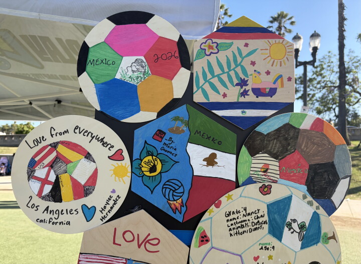 A handful of wooden circles and hexagons, decorated in vibrant colors by children, are affixed to a temporary display board at a park. Palm trees and street lamps cans be seen in the background. 