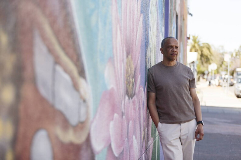 A man wearing light colored pants and a brown shirts stands on a sidewalk, leaning on a building painted with a pastel colored mural.