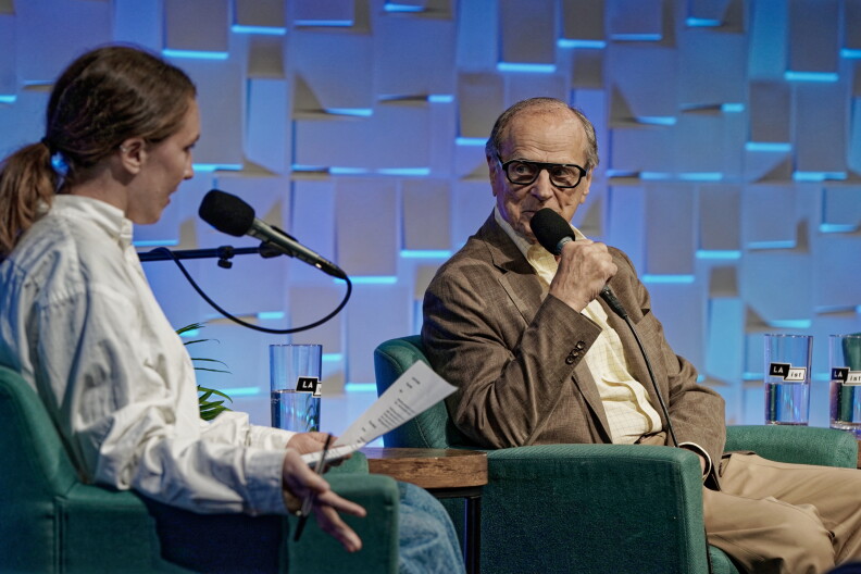 A light-skinned man is holding a microphone and looking to his right. A woman on his right is also speaking behind a microphone and is wearing a white-collared shirt.