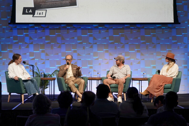 Four people sit on stage. From left to right:  a woman wearing a white shirt, a man wearing glasses and a gray suit, a man wearing a gray shirt and baseball cap, and a woman wearing a brown hat and white shirt. All four hold mics.